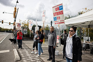 A group of workers stands on a sidewalk holding signs that read "On Strike at Boeing" during a labor protest. The signs also display "Aerospace Machinists" and "Unfair Labor Practices." Some of the protesters wear jackets and sunglasses, and one person holds a dog. A white tent with supplies is set up behind them, and the scene takes place near a road with a traffic light visible in the background under a cloudy sky.