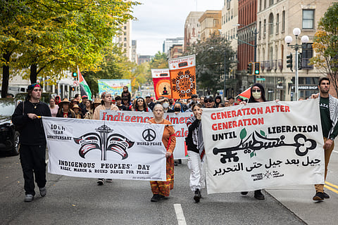 A group of marchers walk down a city street, holding large banners. One banner reads "Indigenous Peoples' Day" and another, in both English and Arabic, says "Generation After Generation Until Liberation." People in the crowd hold additional signs, including one for "Every Child Matters."