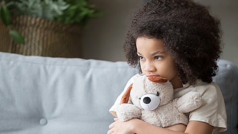 A child who looks a little lonely and sad sits on a couch hugging a stuffed animal and looking off into the distance.