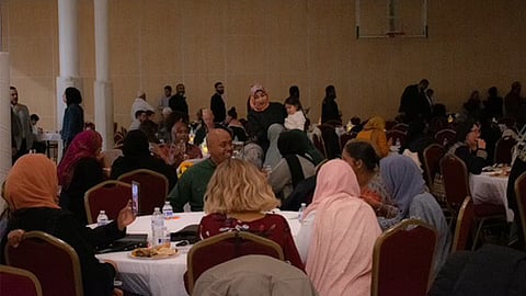 A large gathering of people seated at round tables covered with white tablecloths in a well-lit banquet hall. Attendees, dressed in various styles, including hijabs, are eating, talking, and engaging with one another. A woman in the background is holding a child. A basketball hoop is visible on the far wall.