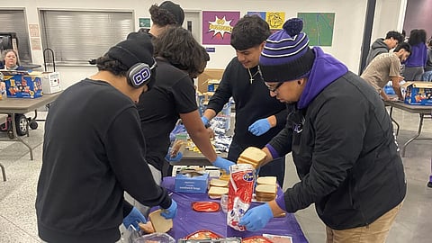 A group of young people assembling sandwiches at a table covered with bread, sandwich bags, and other supplies. They wear gloves and casual clothing, with some sporting beanies. Others in the background are working at additional tables.