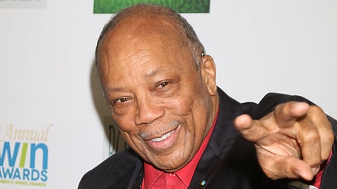 Smiling older man with short hair and a mustache, wearing a dark suit and a red shirt, pointing toward the camera. The background features blurred text and a logo from an event.