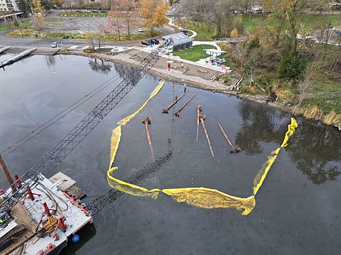 A bird's-eye-view of a shoreline.  A crane arm hovers over logs in a yellow barricaded area in the water.  A parking lot is in the distance.