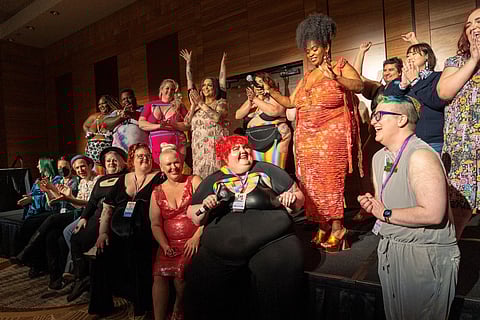 A diverse group of people on a stage, celebrating inclusivity and body positivity. They are smiling, clapping, and cheering, dressed in colorful, expressive outfits. The group includes individuals of varying body types.