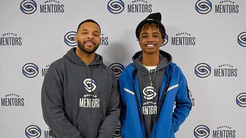 Romell Witherspoon, left, director of mentorship, and mentee Deprince Charles at Seattle Mentors' "Igniting Your Potential" event on Jan. 11. They're standing shoulder to shoulder, Romell standing with his hands clasped in front of himself, and Deprince standing with his hands in his pockets, both of them smiling and in front of a background with the Seattle Mentors logo repeated across it. 