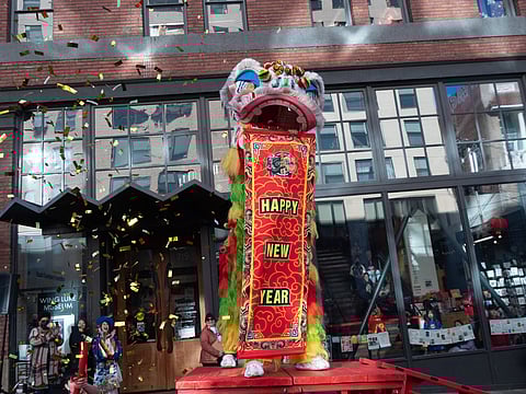 A vibrant Chinese lion dance performance in front of a brick building with large windows, featuring a colorful lion costume holding a red banner that reads "Happy New Year." Gold confetti fills the air as spectators watch from the street and windows. The Wing Luke Museum entrance is visible in the background.