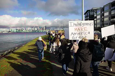 A group of protesters walking along a waterfront path with the city skyline visible in the distance under a cloudy blue sky. A person in the foreground holds a sign that reads, "Immigrants MAKE America Great."