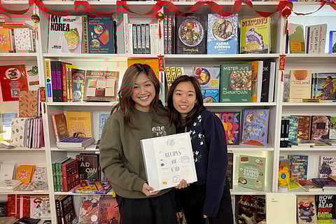 Two smiling Asian-presenting women stand in front of a bookshelf filled with colorful cookbooks and other literary works. They hold a white binder labeled 'Recipes of C-ID,' decorated with hand-drawn food illustrations.