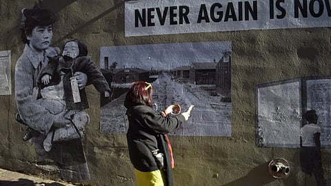A person wearing a black jacket, yellow pants, and a colorful scarf holds a small bundle of sage near a mural painted on a wall. The mural features black-and-white historical images, including a woman carrying a child, a row of barracks, and a sign reading "NEVER AGAIN IS NOW."