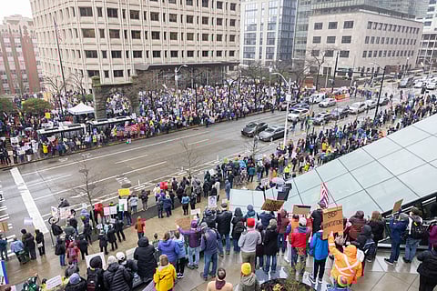 A large protest is captured from an elevated perspective in a city, showing hundreds of demonstrators lining both sides of a wide street. The crowd holds numerous signs with political messages, some reading "STOP DEPORTATIONS" and "NO OLIGARCHY."