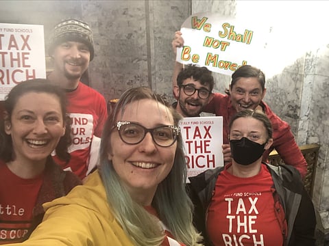Group of smiling activists pose for a selfie inside a marble-walled building, wearing red shirts with messages like “Tax the Rich” and “Fund Our Schools,” while one person holds a colorful sign reading “We Shall Not Be Moved.”