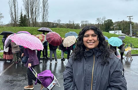 A person with long dark hair smiles at the camera in front of a line of people in the rain with their umbrellas.