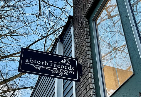 Storefront sign for “absorb records” mounted on the exterior of a brick building. The black sign features white decorative branches and text, with leafless tree branches and a reflection of the sky visible in the window nearby.