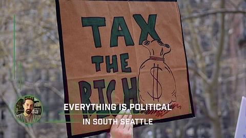 Protest sign reading “TAX THE RICH” in bold green letters with a drawing of a money bag, held up at a demonstration, overlaid with the text “Everything Is Political in South Seattle” and a circular photo of a man in the bottom left corner.