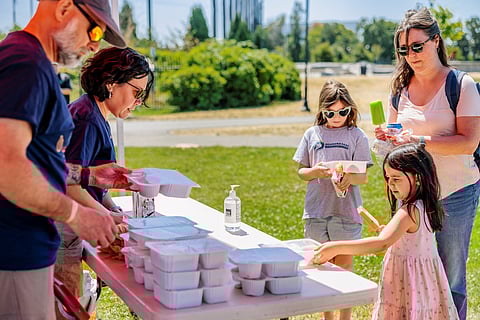 Children and adults receive pre-packaged meals at an outdoor distribution table, with trays stacked and volunteers serving. One child reaches for a meal tray while others enjoy frozen treats.