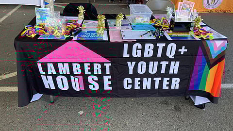 A resource table for Lambert House LGBTQ+ Youth Center, decorated with rainbow wristbands, pamphlets, and promotional materials on a black tablecloth featuring the Lambert House logo and Progress Pride flag colors.