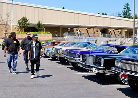 Lowrider car show with classic custom vehicles lined up outside Chief Sealth International High School in Seattle, with attendees walking past.