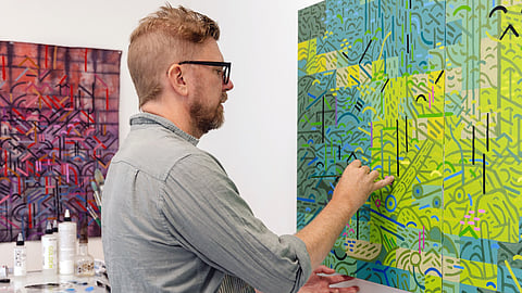 Artist painting detailed geometric abstract artwork in studio, vibrant green and blue patterns, with art supplies and another colorful piece on wall at J Rinehart Gallery.