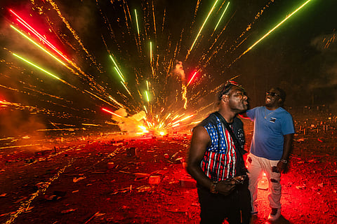 Two men celebrating at Muckleshoot Fireworks Mall at night, standing in front of a large, colorful fireworks explosion; one wearing an American flag shirt and the other in blue and white, with bright red and yellow sparks filling the sky.