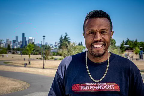 Adonis Ducksworth smiling in a Seattle park with skateboard, wearing a "Built to Grind Independent" T-shirt, Seattle city skyline in background on sunny day.