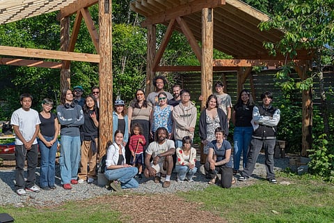 A diverse group of people poses in front of a wooden pavilion structure at the yəhaw̓ Indigenous Creatives Collective.