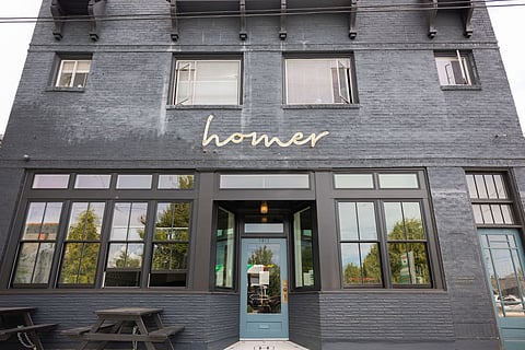 Front exterior of Homer Restaurant in Seattle, showing its dark gray brick facade, large windows, a light blue door, and the "homer" sign above the entrance with outdoor picnic tables in front.