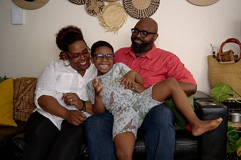 Smiling Black family sitting on a couch at home, with a laughing child in dinosaur pajamas lying across the laps of two joyful parents, framed by woven wall art and cozy decor.