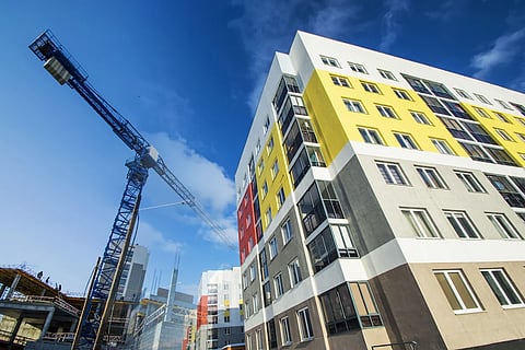 Modern affordable housing complex under construction with colorful exterior panels and a tall blue crane against a bright blue sky.