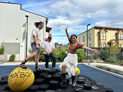 Three Southeast Asian people pose playfully on a circular black playground structure in a sunny urban park in Seattle, with one standing atop a yellow sphere.