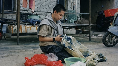 Person seated on a concrete floor engaged in traditional Chinese batik work, dyeing fabric with blue patterns beside plastic bags and buckets, in an outdoor domestic courtyard.