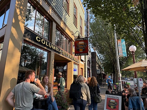 People gather outside Columbia City Ale House on a tree-lined street in Seattle, with outdoor seating, signage for traditional ales and food, and Columbia City banners visible along the sidewalk.
