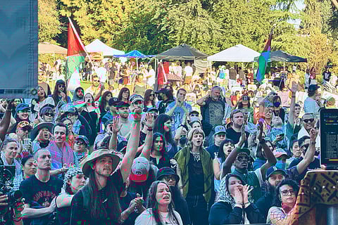 Large, diverse crowd at an outdoor event waving Palestinian flags and wearing keffiyehs, with people cheering and raising their hands under tents and trees.