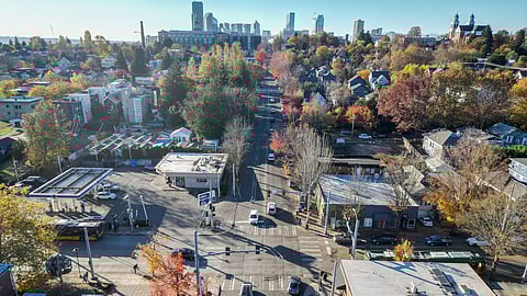 Aerial view of a busy Seattle intersection with cars, buses, and cyclists, surrounded by shops, trees with autumn colors, and residential neighborhoods, with the downtown Seattle skyline visible in the distance.
