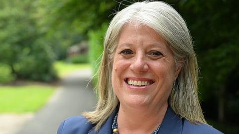 Smiling woman with gray hair wearing a navy blazer and beaded necklace, standing outdoors on a pathway with green trees and grass in the background.