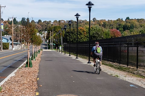 A cyclist rides along a newly paved multi-use trail lined with young trees and black lampposts, with a fence and colorful autumn trees in the background.