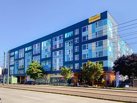 Modern blue and teal mid-rise apartment building in Seattle's Othello neighborhood with "Now Leasing" signs, located along a street with light rail tracks, trees, and parked cars under a clear blue sky.