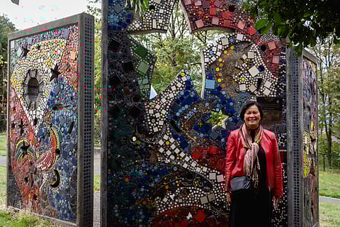 Southeast Asian-presenting woman in a red jacket standing beside a colorful outdoor mosaic art installation made of vibrant tiles in blue, red, yellow, and white patterns at a park.