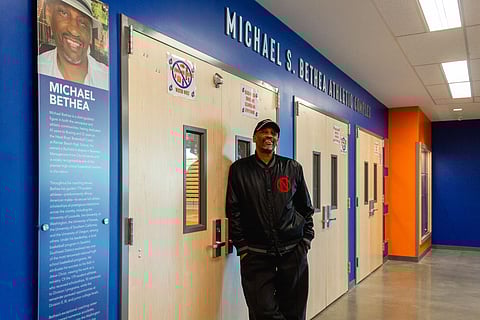 Black-presenting man standing and smiling in a hallway outside the Michael S. Bethea Athletic Complex, next to a blue wall display honoring coach Michael Bethea’s career and achievements in basketball and education.