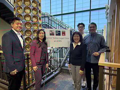 Five people stand indoors beside a sign for the Rainier Valley Legal Clinic at ACRS, with logos for the Asian Bar Association and King County Bar, posing in front of a staircase and decorative gold wall display.
