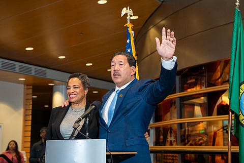 Smiling biracial Black man in a blue suit with his arm raised beside a Black woman at a podium, both standing in front of U.S. and state flags.
