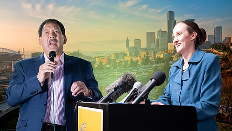 Two people speaking at separate podiums against a backdrop of the Seattle skyline, with multiple microphones in the foreground and warm sunset lighting over the city.