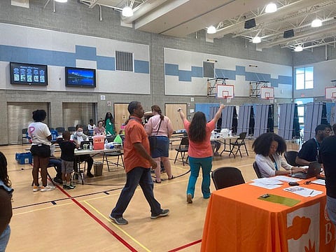 Community event inside a gym with volunteers seated at tables assisting attendees, medical staff working at stations, and people walking through the space beneath bright lights and basketball hoops.