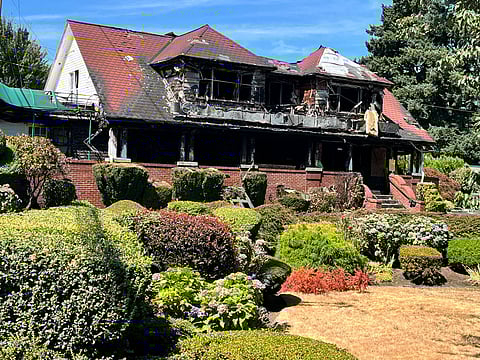 Burned two-story house with a charred roof and blackened walls, showing severe fire damage, surrounded by well-kept shrubs, hedges, and blooming hydrangeas under a bright blue sky.