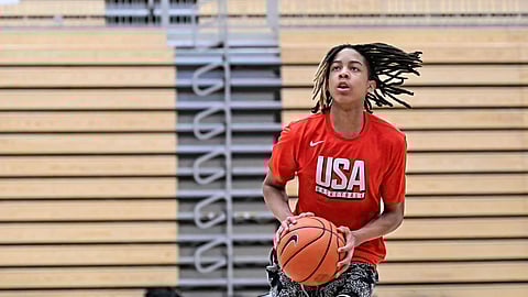 Teen basketball player in a red USA Basketball shirt holds a ball while jumping for a shot in an indoor gym, wooden bleachers blurred behind him.