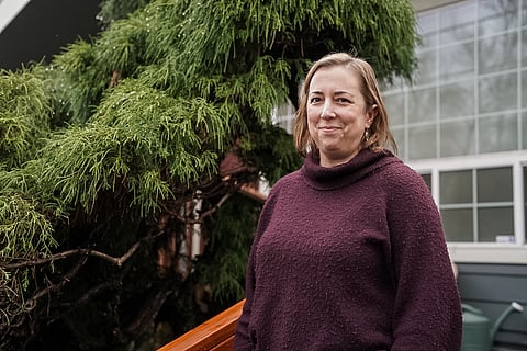 A female-presenting figure, with shoulder-length hair and a maroon cowl-neck sweater, stands on a staircase outside of a house. A tree is in the background.