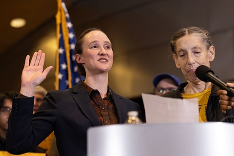 Newly sworn-in Seattle mayor raises her right hand while taking the oath at a podium, with an American flag behind her and another person reading from a paper during the inauguration ceremony.