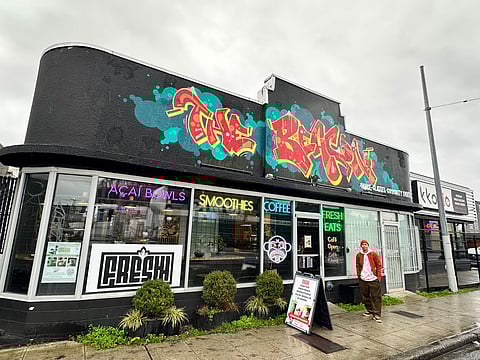 Brysen Angeles stands on the sidewalk outside The Beacon studio on Rainier Avenue South, neon window signs, and café storefront visible.