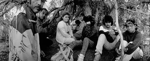 Black-and-white photo of a group of migrants sitting under trees near Interstate 5 at the U.S.–Mexican border in San Diego County, California, 1989, appearing tired and watchful.