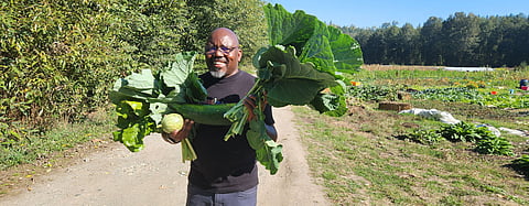 David Bulindah at Horseneck Farm with crops grown by Wakulima USA farmers during the 2025 season. He's smiling and holding the large crops in his hand; the farm is visible behind him.