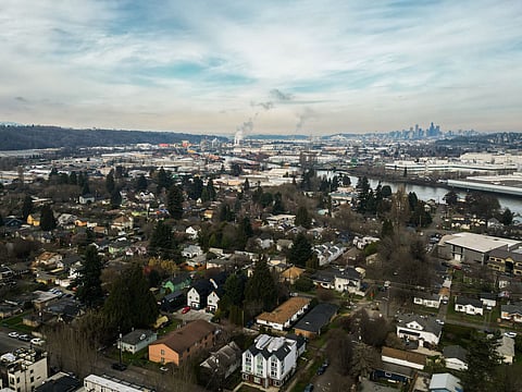 Aerial view of Seattle’s South Park neighborhood with homes and trees in the foreground, industrial buildings along the Duwamish River, and steam rising from the Ash Grove Cement plant under a cloudy sky.